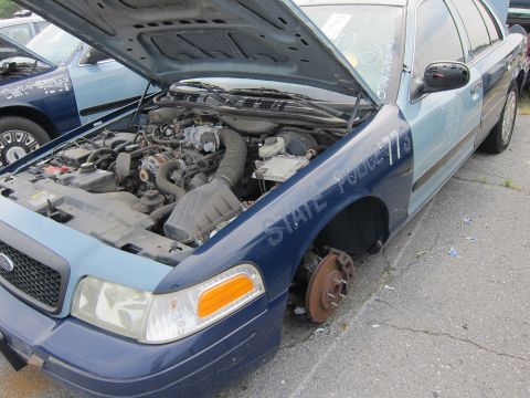 2001 Ford Crown Victoria - A damaged 2001 Ford Crown Victoria police car is shown with its hood open and a missing front wheel, revealing the engine compartment.