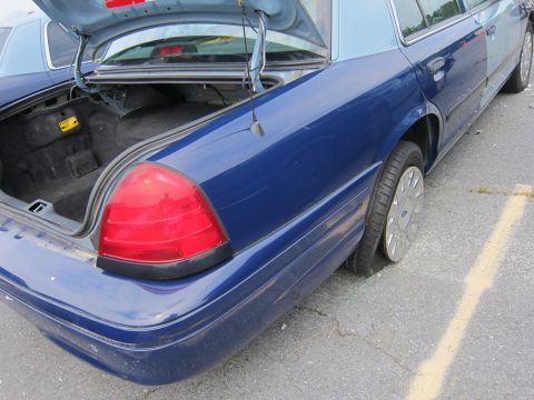 2001 Ford Crown Victoria - The trunk of a blue sedan is open, revealing an empty cargo area and a flat tire on the rear passenger side.