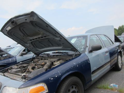 2001 Ford Crown Victoria - A two-tone sedan sits in a junkyard with its hood and trunk open, revealing the engine and interior. Writing is visible on the windshield.
