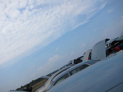 2001 Ford Crown Victoria - Multiple vehicles are parked in a junkyard under a bright, cloudy sky.