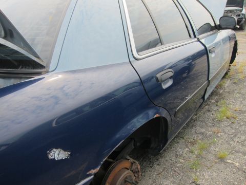 2001 Ford Crown Victoria - A blue 2001 Ford Crown Victoria sits in a junkyard with its wheels removed and trunk open.