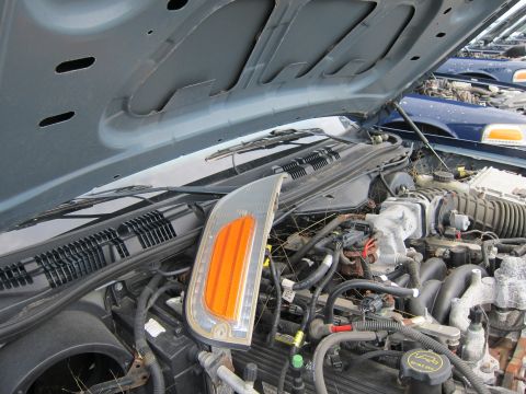2001 Ford Crown Victoria - Engine bay of a car with a detached headlight assembly. Other vehicles are parked in the background.