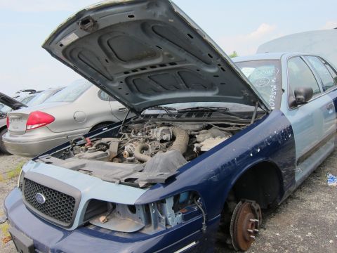2001 Ford Crown Victoria - A two-tone Ford Crown Victoria sits in a junkyard with its hood open, revealing the engine compartment and missing front fender.