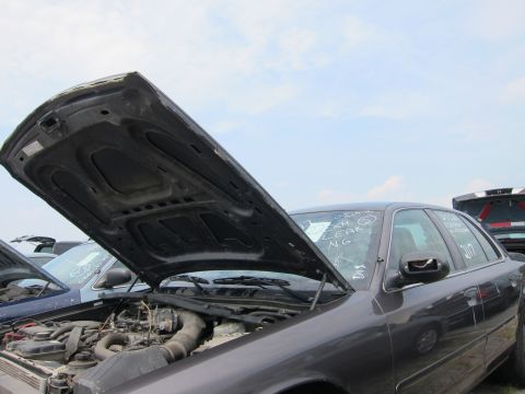 2001 Ford Crown Victoria - A dark sedan sits in a junkyard with its hood open, revealing the engine compartment. Other vehicles are visible in the background.