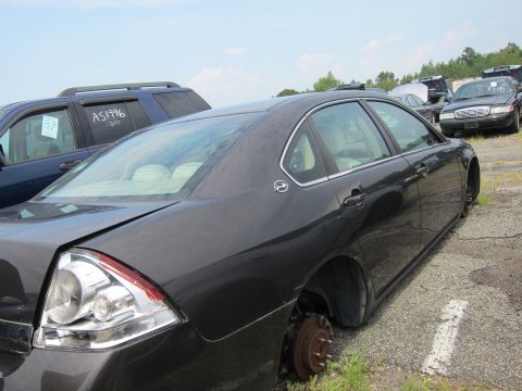 2001 Ford Crown Victoria - A dark sedan sits in a junkyard with its wheels removed, parked next to other vehicles under a cloudy sky.