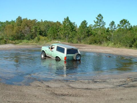 1994 Ford Bronco - A light green SUV is partially submerged in water on a dirt road, with trees in the background.