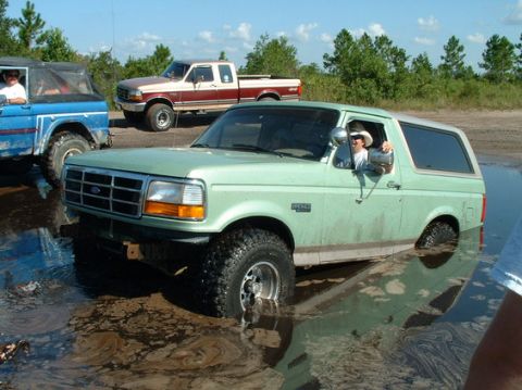 1994 Ford Bronco - A light green 1994 Ford Bronco is stuck in a muddy puddle, with another Bronco and a pickup truck visible in the background.