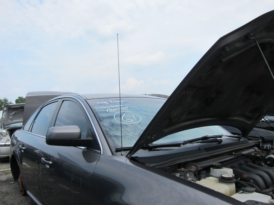 Dark sedan in a junkyard, hood open, showing engine. Writing on windshield says 'Eng light - Hesitate Auction 61'. Another car is visible behind it