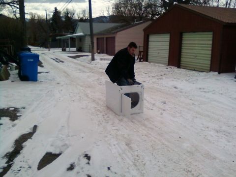 2006 Ford E350 - A man sits in a white appliance, possibly a dryer, on a snow-covered road in front of houses and garages.