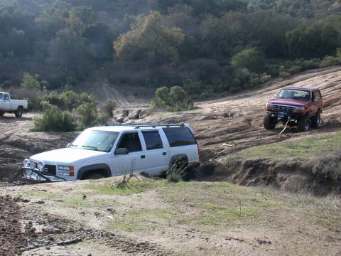 1996 Ford Bronco - A red Ford Bronco is being pulled up a muddy incline by a winch cable, while a white SUV is stuck in the mud below.