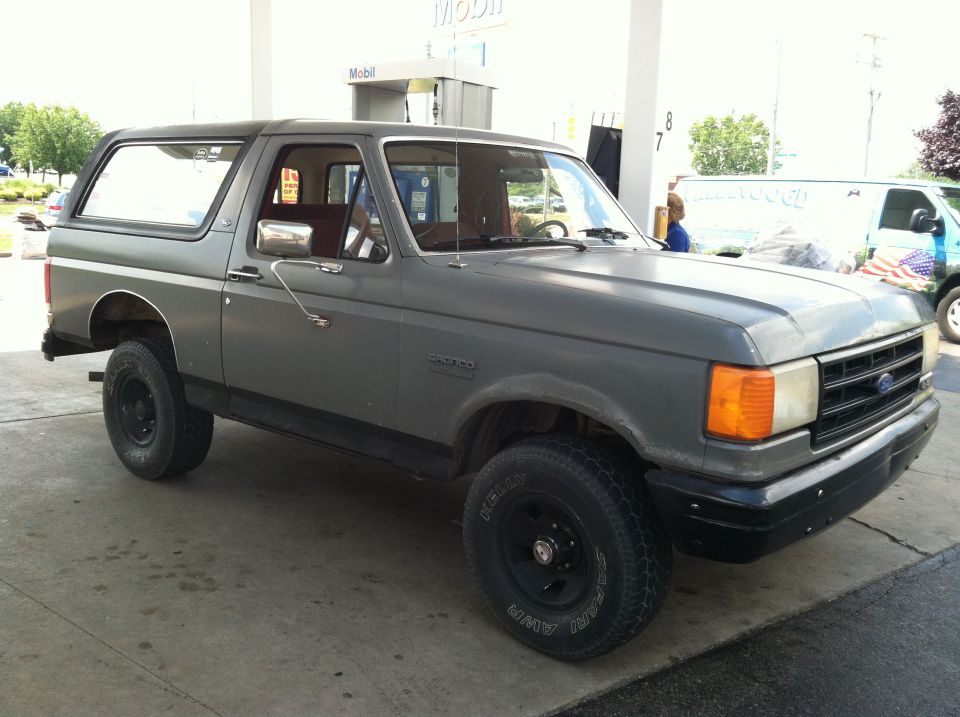 Gray 1987 Ford Bronco parked at a gas station with a white van in the background