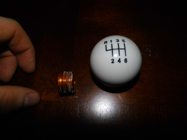 White shift knob showing a 6-speed manual transmission pattern (R, 1, 3, 5 top; 4, 2, 6 bottom) next to a small copper coil. A hand is visible on the left