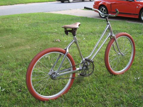 Chrome bicycle with red tires and brown seat on grass, red car in background