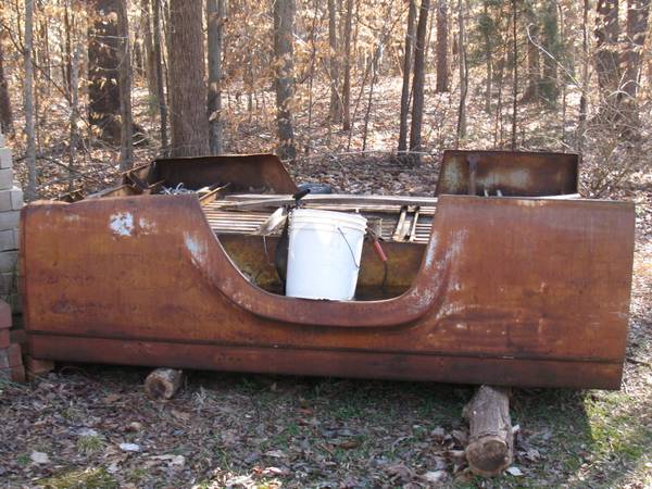 Rusted truck bed with a white bucket inside, resting on logs in a wooded area