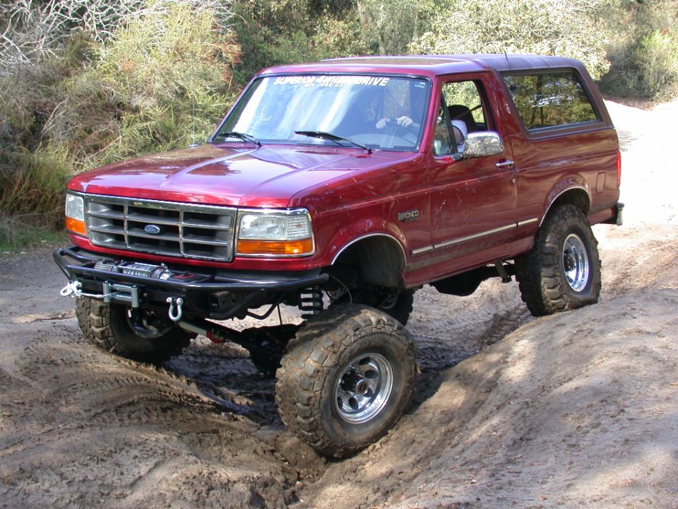 Red lifted Ford Bronco flexing on a dirt trail with winch bumper and polished wheels