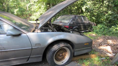 1987 Pontiac Firebird - Two cars sit in an outdoor setting; one is a 1987 Pontiac Firebird with its hood up.