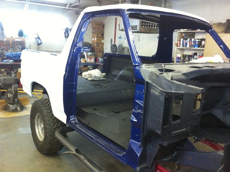 Truck body being worked on in a garage. White cab, blue doors, and bare metal chassis visible