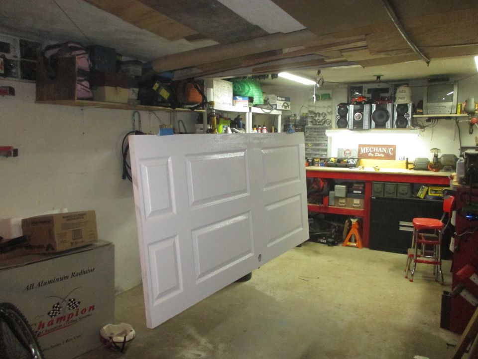 Garage interior with a white paneled door leaning against a wall, shelves with items, and a workbench