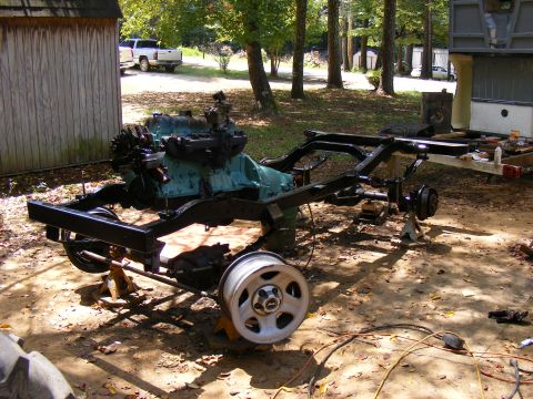 Vehicle frame and engine on jacks, with a white pickup truck in the background