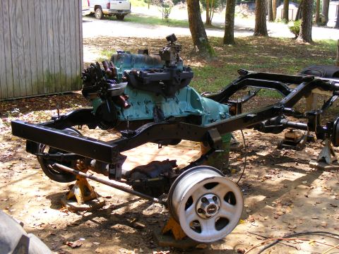 Jeep frame on jack stands with engine and one wheel attached. A white pickup truck is visible in the backgroun
