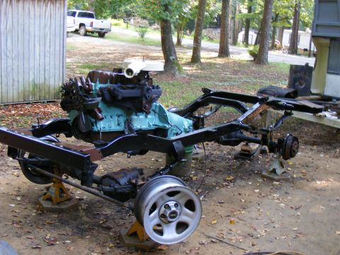 Vehicle frame and engine assembly on jack stands outdoors. A white pickup truck is visible in the background