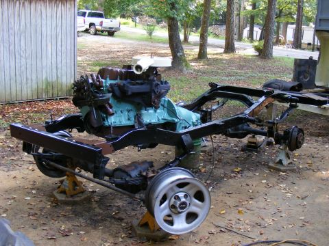 Vehicle frame and engine on jack stands, with a white pickup truck in the background