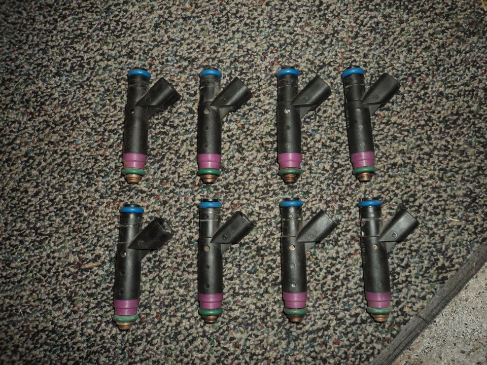 Eight fuel injectors with blue tops and purple bands on a carpeted surface