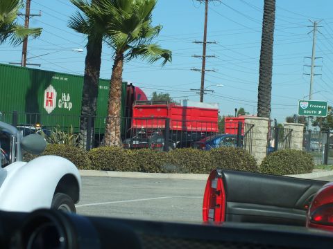 2003 Ford Crown Victoria - Red trucks and a green Hub Group shipping container sit behind palms and a fence, a freeway 'SOUTH' sign visible, shot over a car.
