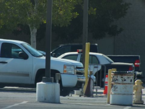 2003 Ford Crown Victoria - white pickup truck and a black-and-white police sedan sit beside concrete bollards, a traffic cone, and a rusty barrel.