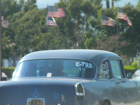 2003 Ford Crown Victoria - gray 1950s-style sedan marked 'C-793' on its windshield rolls past a backdrop of American flags and trees.