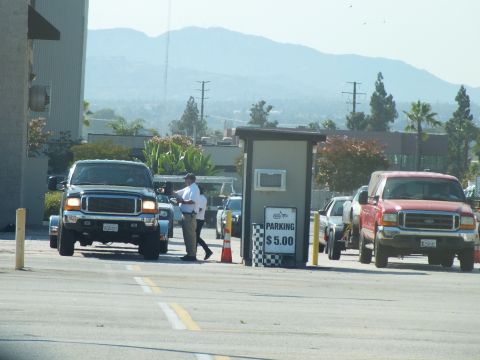 2003 Ford Crown Victoria - black Ford Super Duty and a red Ford pickup flank a checkered '$5.00 parking' booth with an attendant, mountains in the haze.