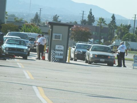 2003 Ford Crown Victoria - vintage Mustang and a police-style sedan queue at a checkered '$5.00 parking' booth where attendants direct traffic.