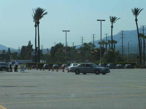 2003 Ford Crown Victoria - gray four-door sedan drives across a wide empty parking lot ringed by palm trees, flags and distant hills.