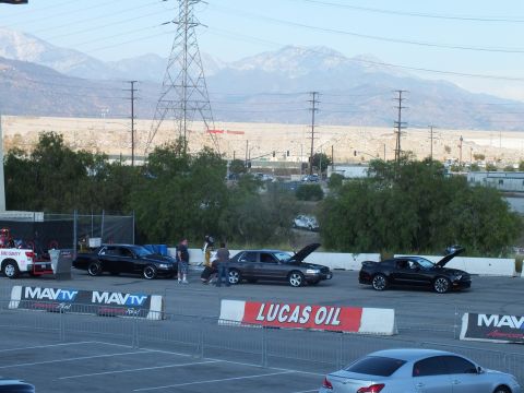 2003 Ford Crown Victoria - Sedans and a Mustang sit with hoods up behind MAVTV and Lucas Oil banners beneath transmission towers and mountains.