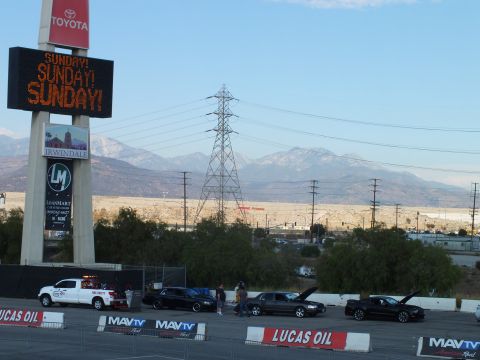 2003 Ford Crown Victoria - pylon sign flashing SUNDAY SUNDAY SUNDAY rises above staged cars and Lucas Oil banners with snowy mountains behind.
