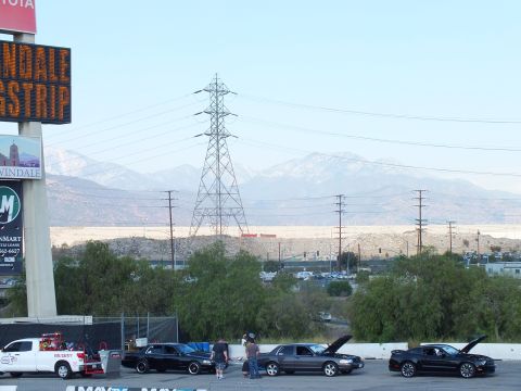 2003 Ford Crown Victoria - Sedans and a Mustang stage with hoods up beneath an Irwindale Dragstrip sign, transmission towers and mountains behind.