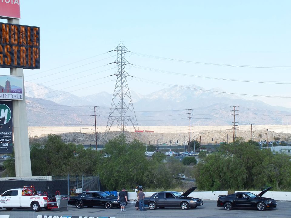 Cars with open hoods staged below an Irwindale Dragstrip sign with power towers and mountains