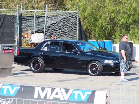 2003 Ford Crown Victoria - blacked-out Mercury sedan on dark wheels, marked with race numbers, idles by a spectator behind a MAVTV banner.