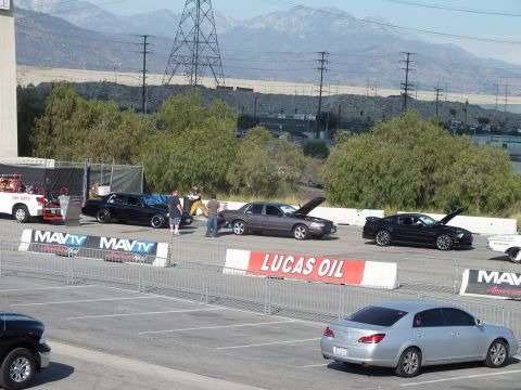 2003 Ford Crown Victoria - From above, sedans and a Mustang stage with hoods open behind Lucas Oil and MAVTV banners with mountains beyond.