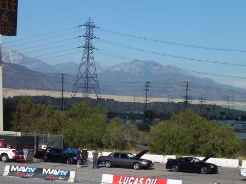 2003 Ford Crown Victoria - Staged sedans and a Mustang line up below a large transmission tower and snow-dusted mountains behind event banners.