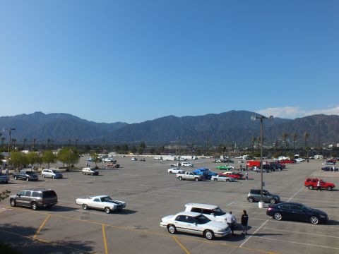 2003 Ford Crown Victoria - From above, classic coupes and modern sedans are parked across a wide lot with palm trees and mountains behind.