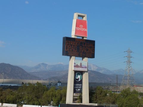 2003 Ford Crown Victoria - tall Toyota pylon displays STREET LEGAL DRAG RACING in LED over an Irwindale event lot with mountains behind.
