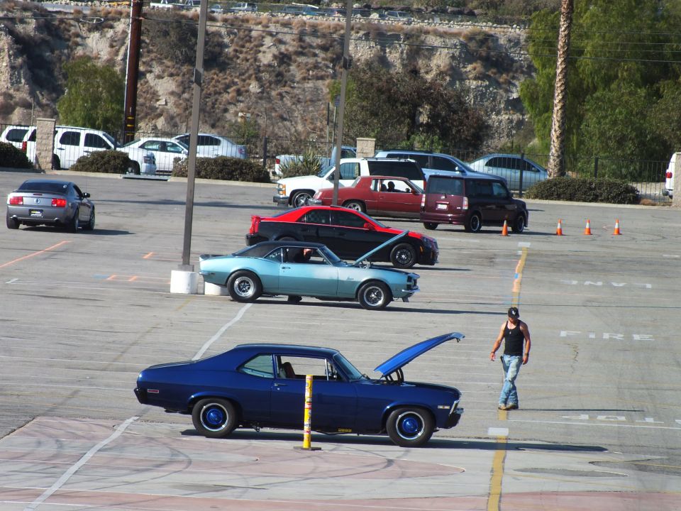 Blue classic coupe with open hood and a turquoise Camaro staged in a lot, man walking by