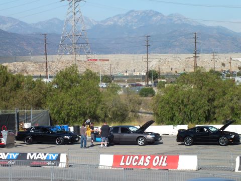 2003 Ford Crown Victoria - Spectators stand among black sedans and a Mustang with hoods open behind a Lucas Oil banner and distant mountains.