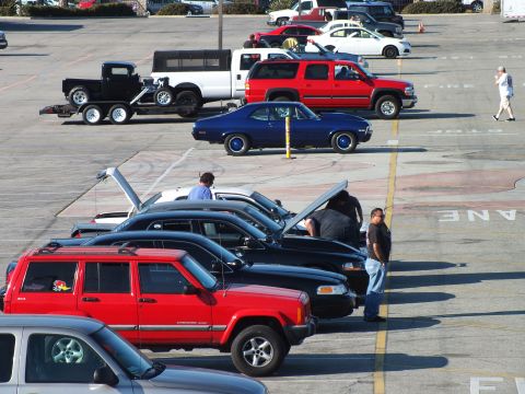 2003 Ford Crown Victoria - long row of vehicles, a red Jeep, blue coupe, hooded sedans and a trailered hot rod, fill an outdoor event lot.
