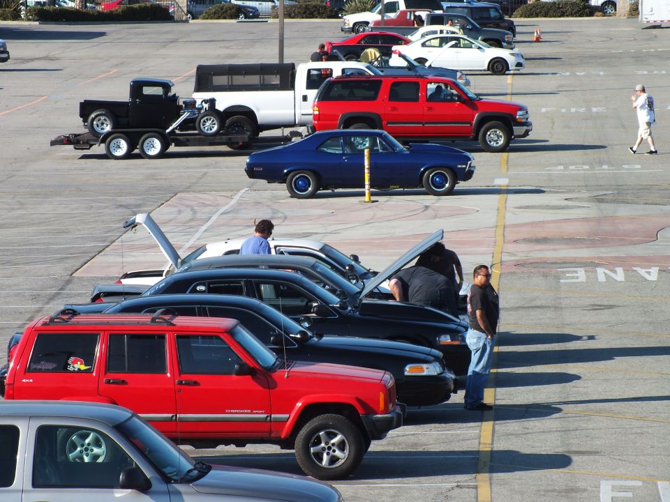 Row of vehicles in a lot: red Jeep, blue coupe, sedans with open hoods and a car trailer