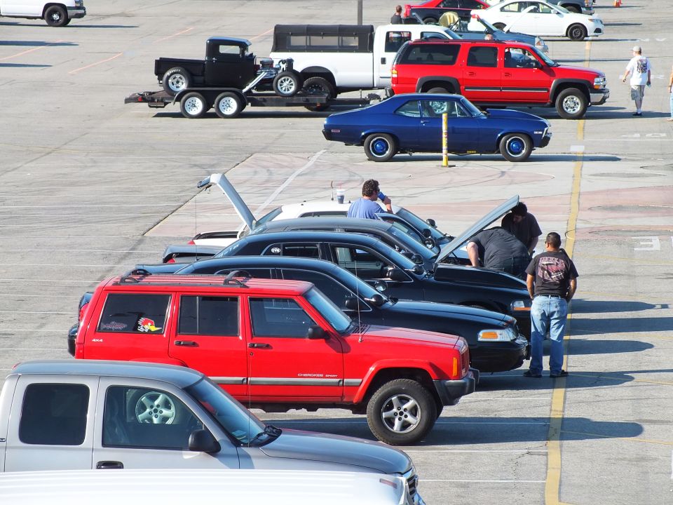 Row of cars with raised hoods, a red Jeep, blue coupe and trailered hot rod in a lot