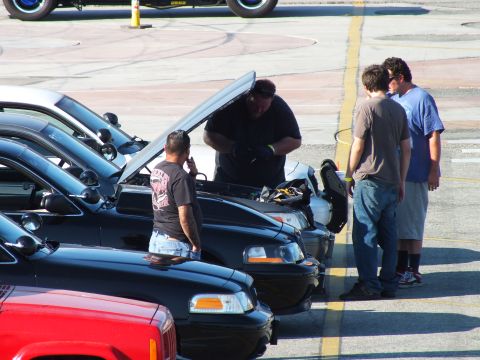 2003 Ford Crown Victoria - Several men inspect the open engine bay of a black car parked in a diagonal lineup on pavement.