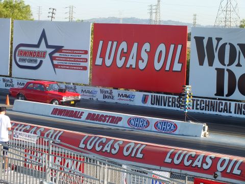 2003 Ford Crown Victoria - red classic car sits at the start of the Irwindale Dragstrip beneath large Lucas Oil and event-center banners.