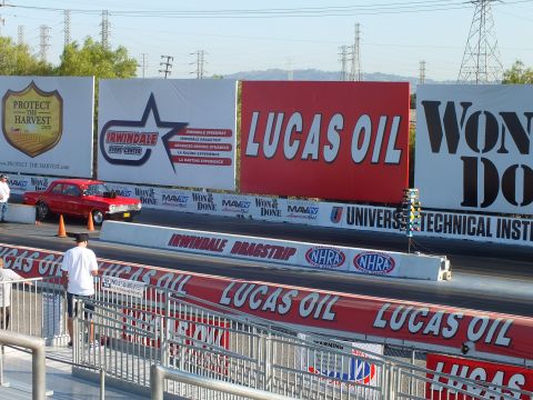 2003 Ford Crown Victoria - red classic car stages on a drag strip lined with Lucas Oil and Protect the Harvest banners under a clear sky.